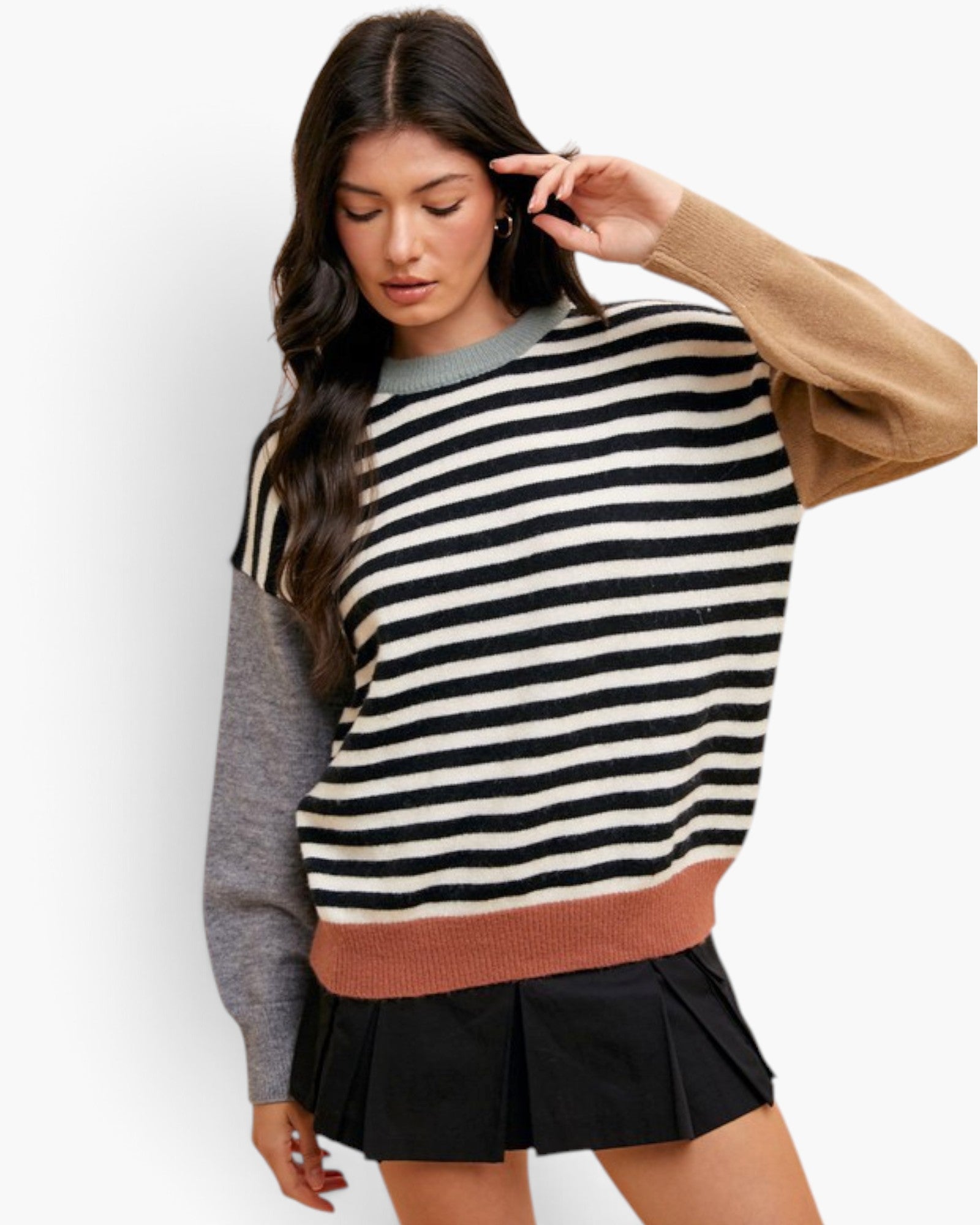 A woman modeling a color-block knit sweater with horizontal black and cream stripes across the chest and sleeves, a solid gray long sleeve, a tan long sleeve, and a dusty rose ribbed hem, paired with a black pleated mini skirt against a light gray studio background.
