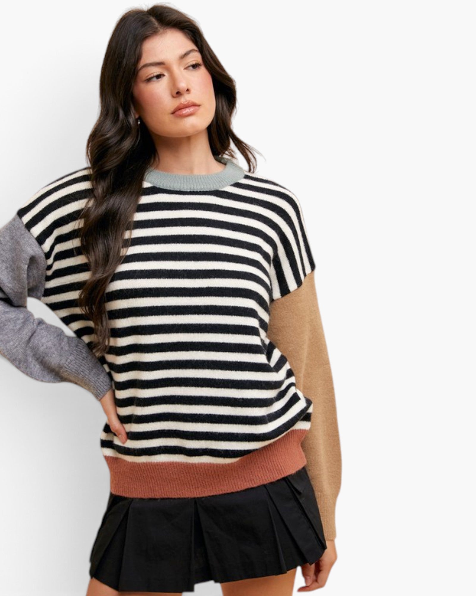 A woman modeling a color-blocked knit sweater with horizontal black and off-white stripes, a gray sleeve, a tan sleeve, and a dusty rose ribbed hem, paired with a black pleated mini skirt against a plain white background.