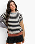A woman modeling a color-blocked knit sweater with horizontal black and off-white stripes, a gray sleeve, a tan sleeve, and a dusty rose ribbed hem, paired with a black pleated mini skirt against a plain white background.