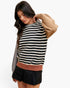 A woman modeling a knit sweater with horizontal black and cream stripes, featuring a solid tan shoulder, a dusty rose ribbed hem, and a gray ribbed collar, paired with a black pleated mini skirt against a light gray background.