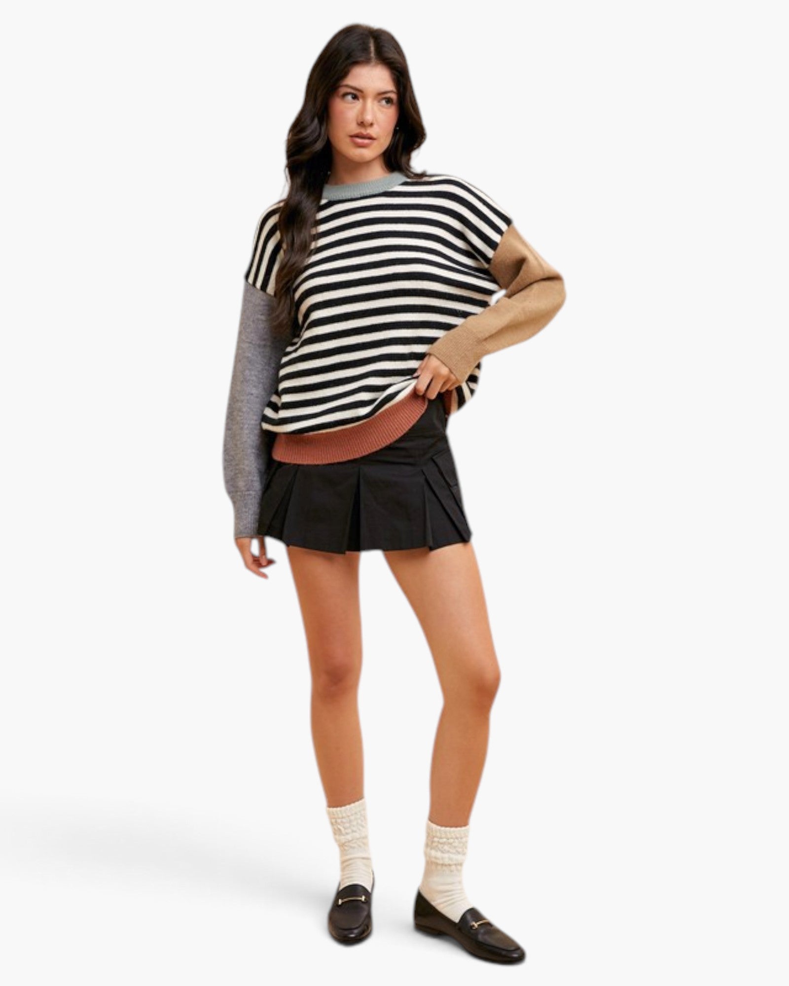 A model wearing a black and white striped knit top with long gray and beige sleeves, a black pleated mini skirt, cream socks, and dark loafers, photographed against a clean white studio background.