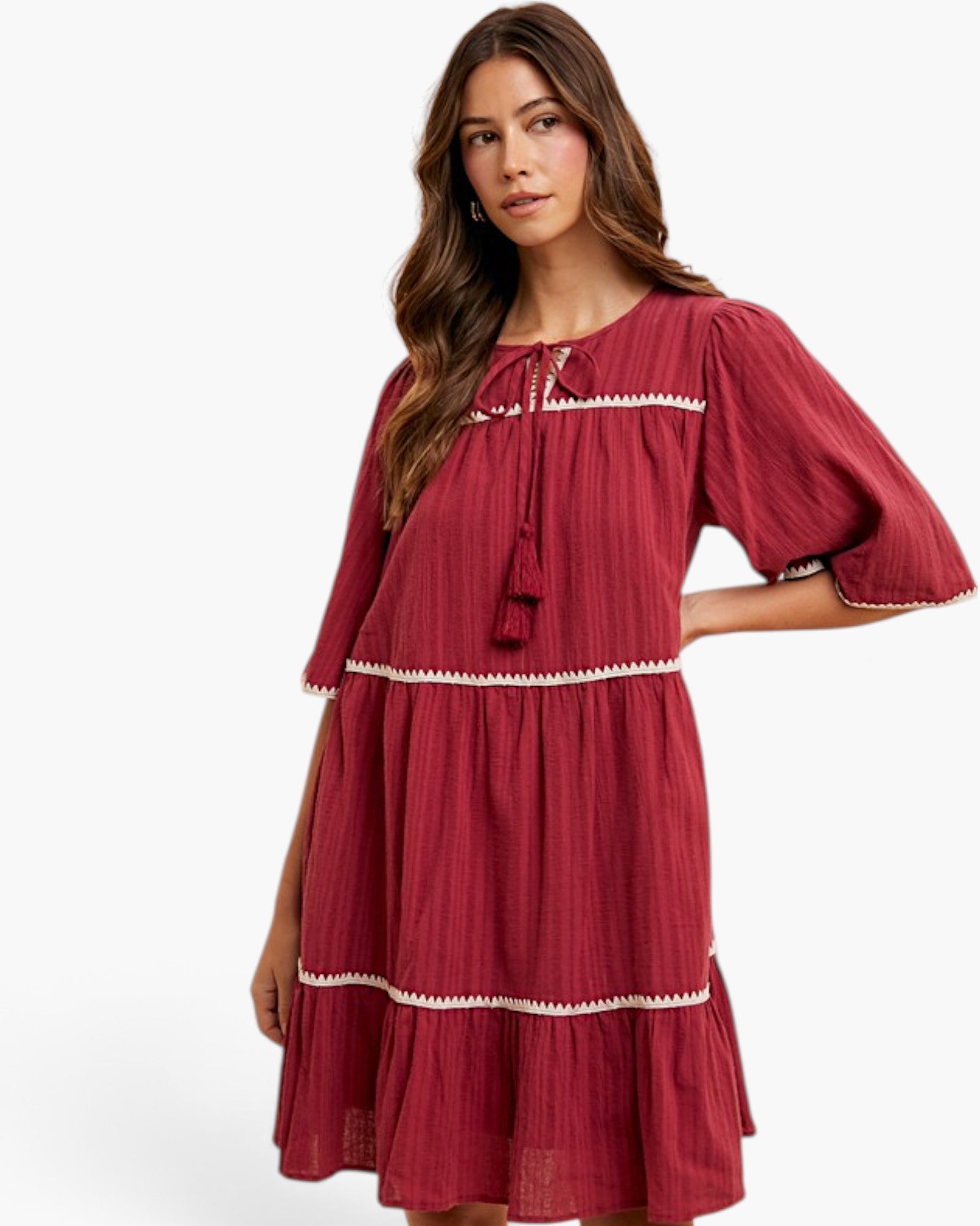 A woman modeling a tiered red dress with white embroidered trim and tassel tie details, photographed against a clean white studio background.