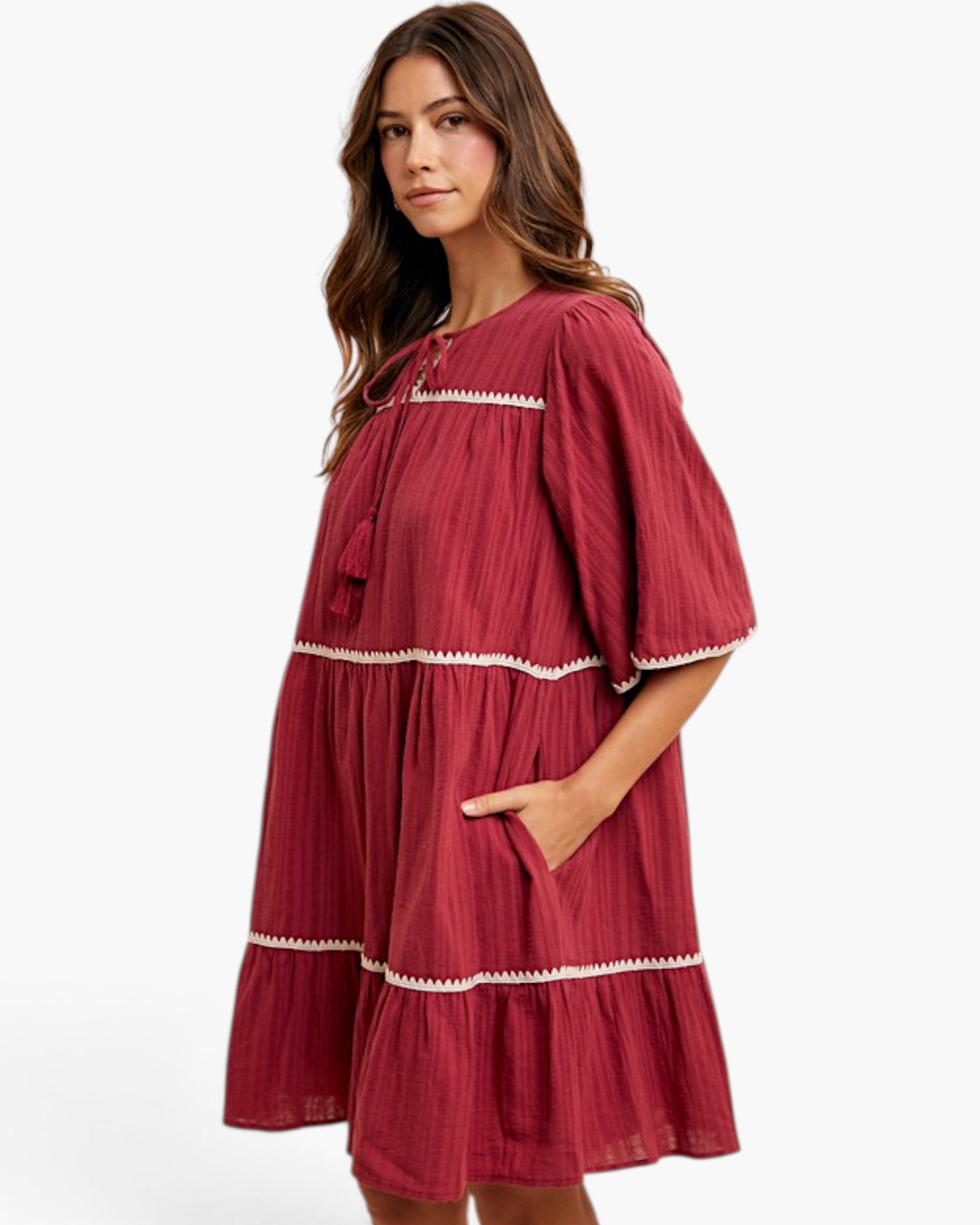 A woman wearing a tiered, short-sleeved dress in a deep burgundy red with white decorative trim, photographed against a plain white studio background.
