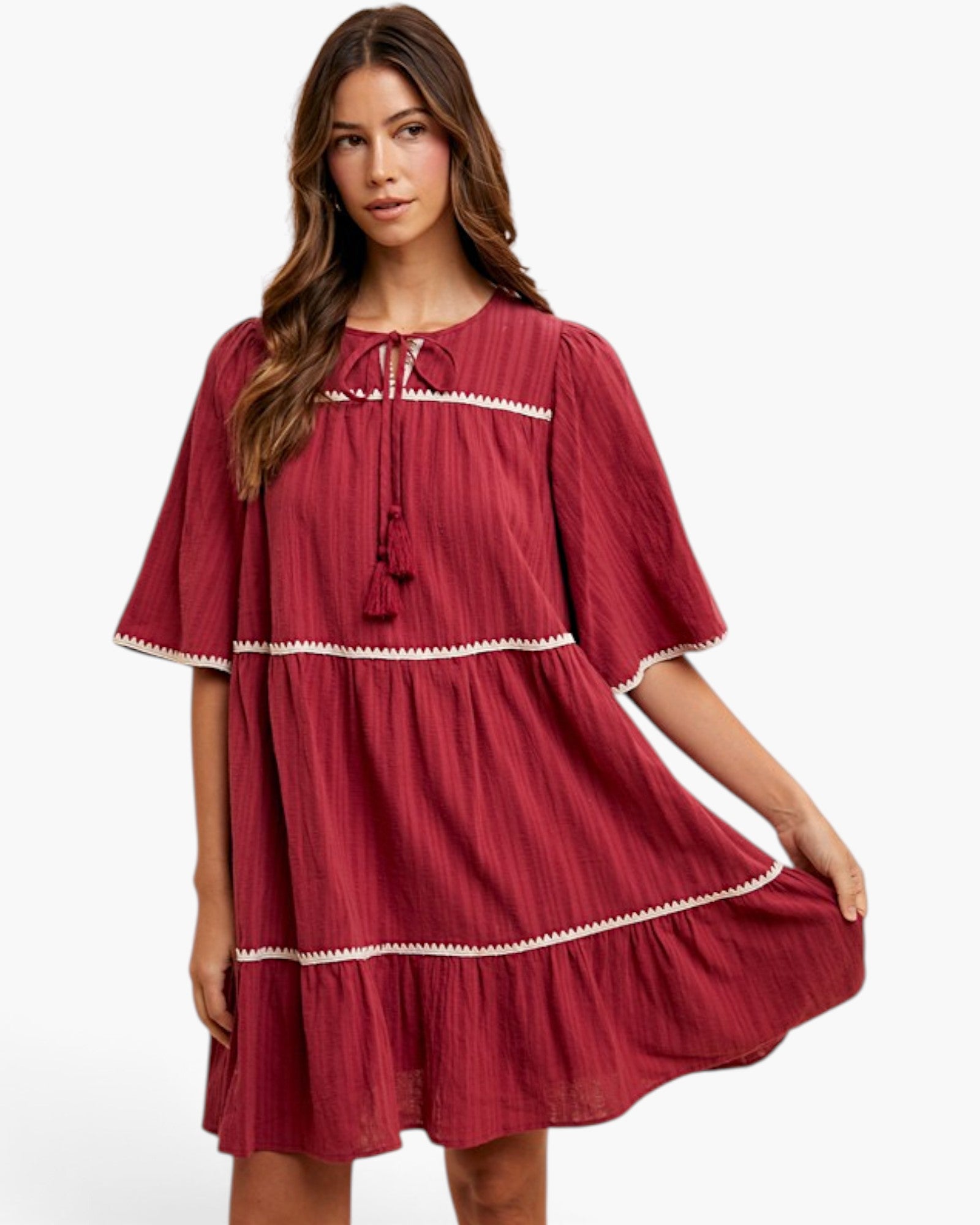 A woman modeling a tiered, short-sleeved dress in a deep red or burgundy crinkle fabric with white embroidered trim details, set against a plain white studio background.