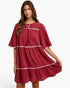 A woman modeling a tiered, short-sleeved dress in a deep red or burgundy crinkle fabric with white embroidered trim details, set against a plain white studio background.