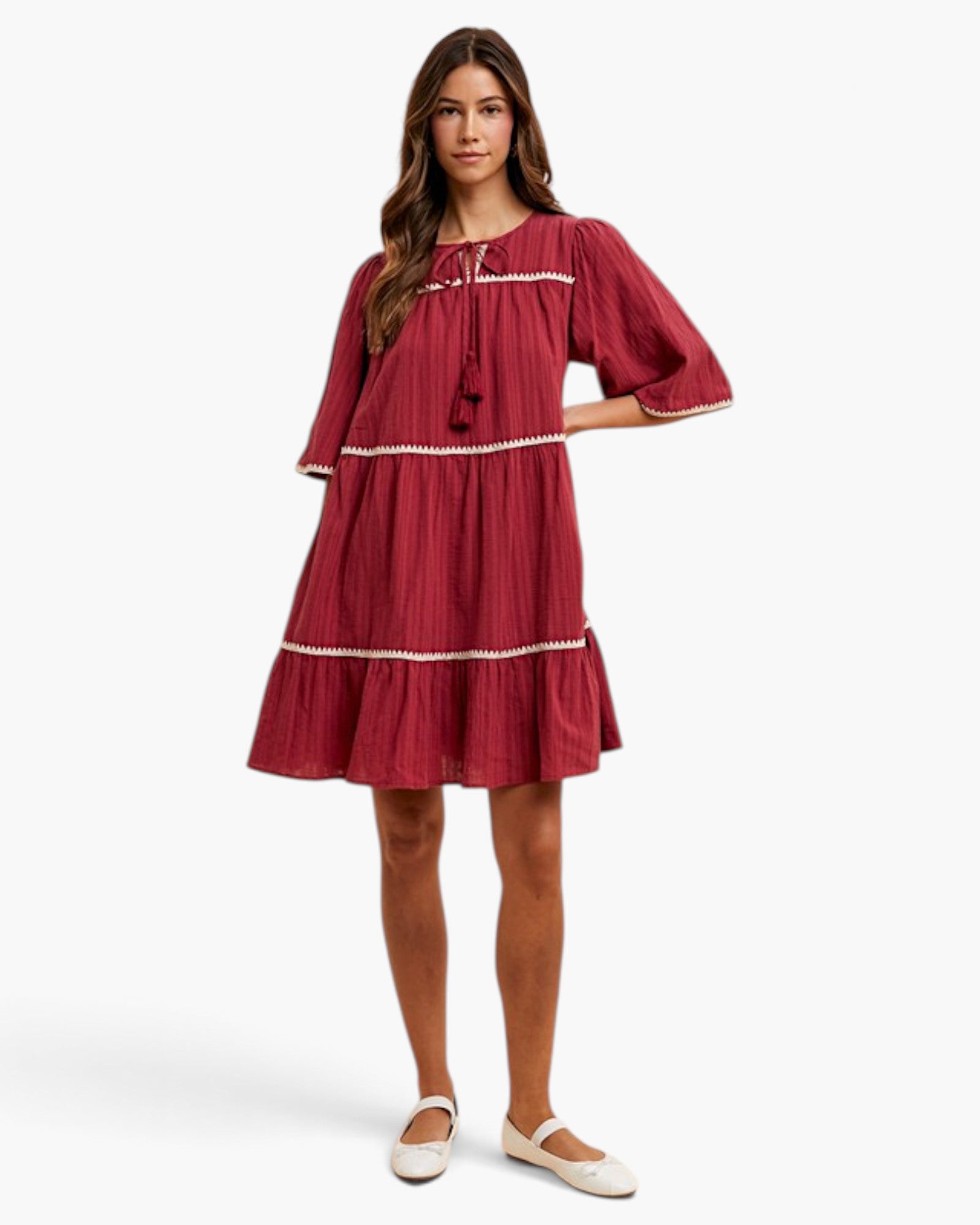 A woman modeling a tiered red dress with white trim and short puff sleeves, paired with white flats, against a light gray studio background.