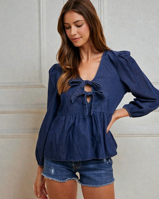 Woman wearing a blue blouse with ruffled details and denim shorts against a white wall.