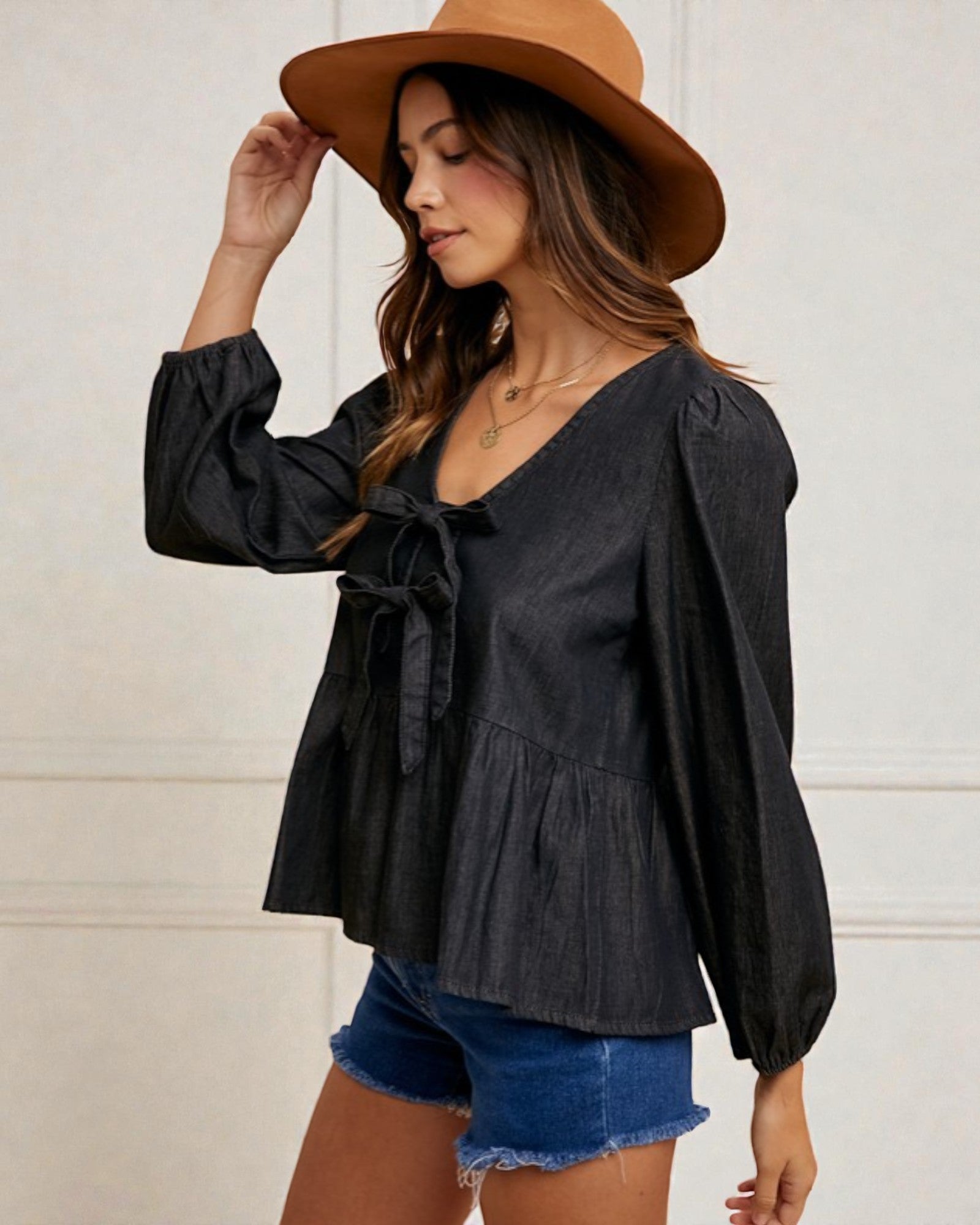 Woman wearing a black blouse with long sleeves and denim shorts, standing against a white wall.