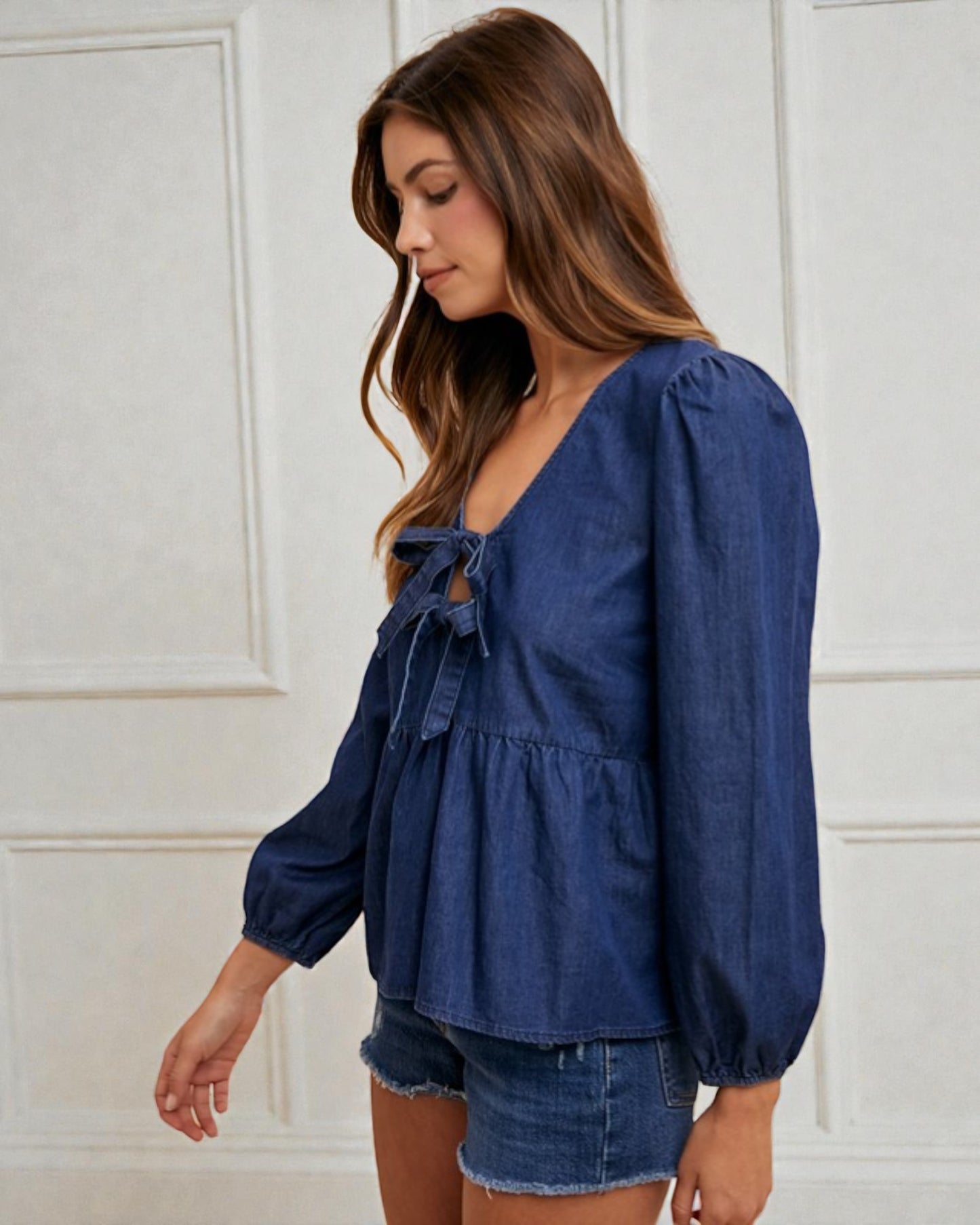 Woman wearing a blue denim top with balloon sleeves against a white paneled wall.