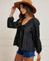 Woman wearing a black blouse with long sleeves and denim shorts, standing against a white wall.