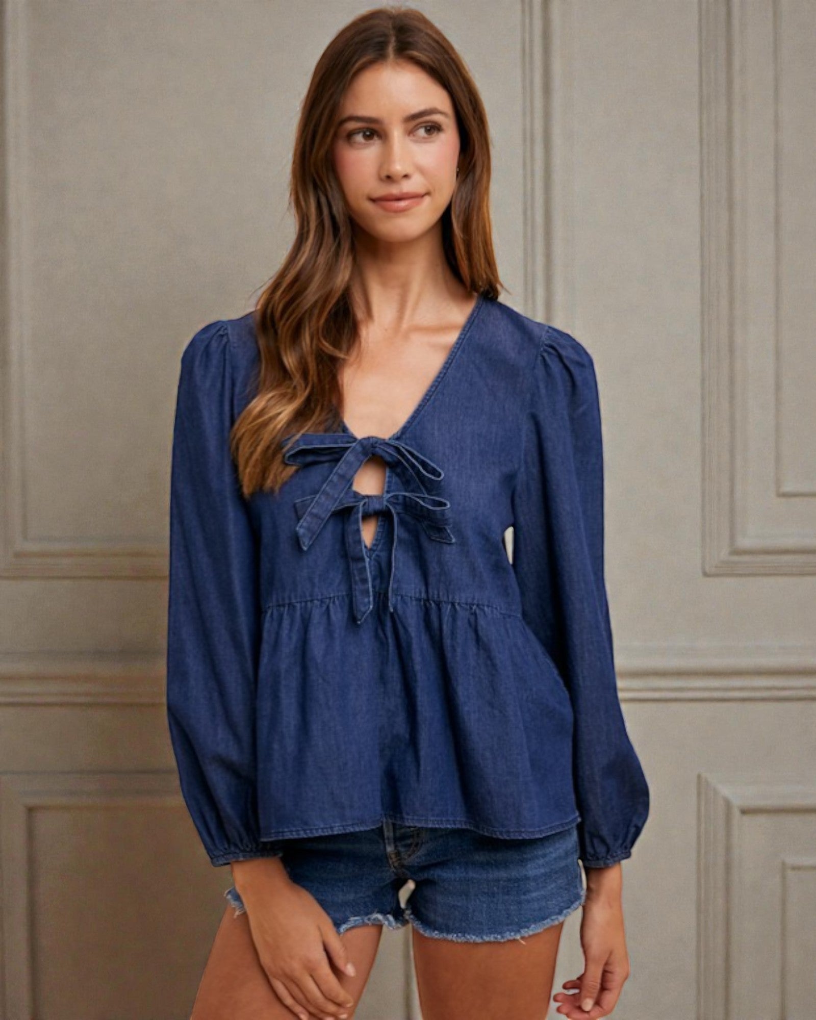 Woman wearing a blue denim top with a bow detail against a neutral wall.