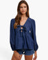 A woman modeling a dark blue denim long-sleeve blouse with a tie-front keyhole detail and ruffled sleeves, paired with denim shorts against a plain white background.