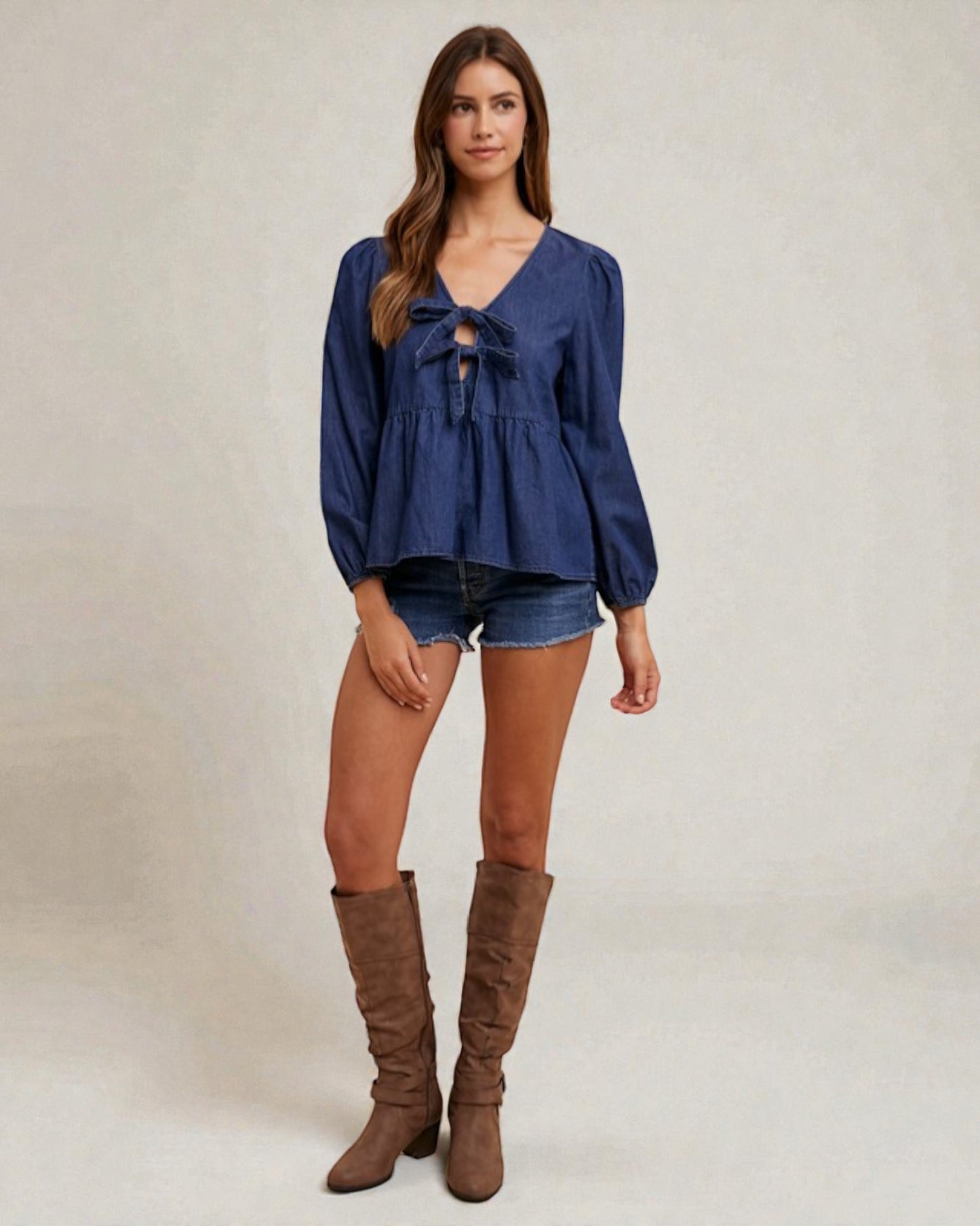 Woman wearing a blue top and denim shorts with brown knee-high boots on a plain background