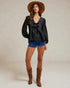 Woman wearing a black blouse, blue shorts, and brown boots with a hat on a plain background