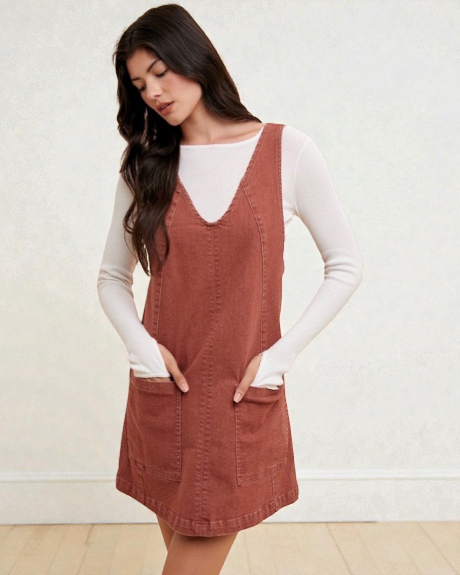 Woman wearing a brown pinafore dress over a white long-sleeve shirt against a white background