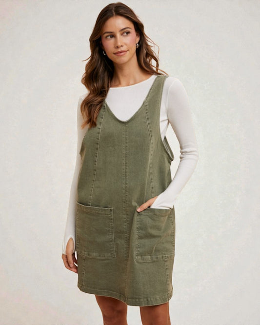 Woman wearing a green denim pinafore dress over a white long-sleeve shirt on a light gray background