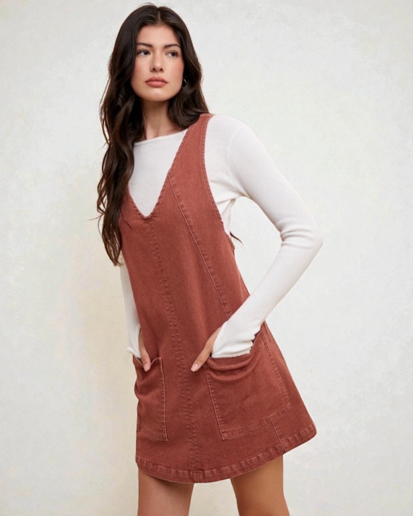 Woman wearing a brown pinafore dress over a white long-sleeve shirt against a plain background