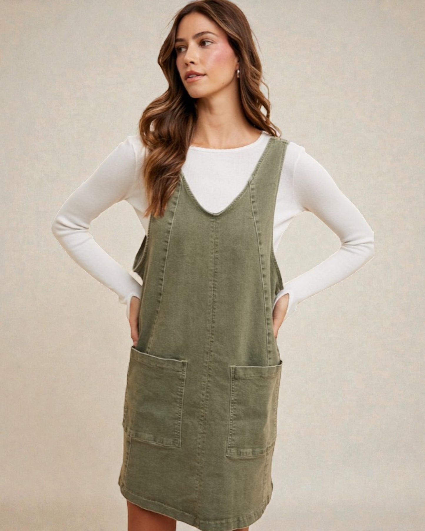 Woman wearing a green pinafore dress over a white long-sleeve shirt against a beige background