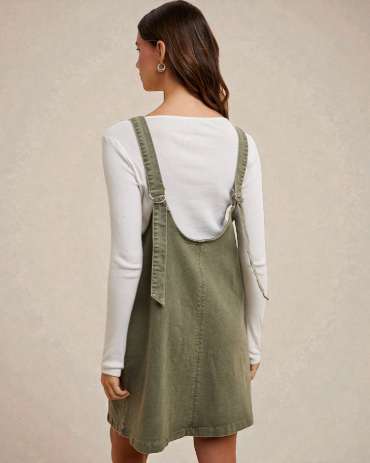 Person wearing a green denim pinafore dress over a white long-sleeve shirt against a beige background