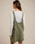 Person wearing a green denim pinafore dress over a white long-sleeve shirt against a beige background