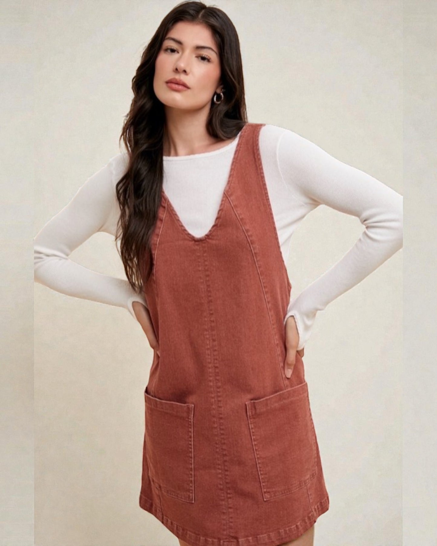 Woman wearing a brown pinafore dress over a white long-sleeve shirt on a beige background