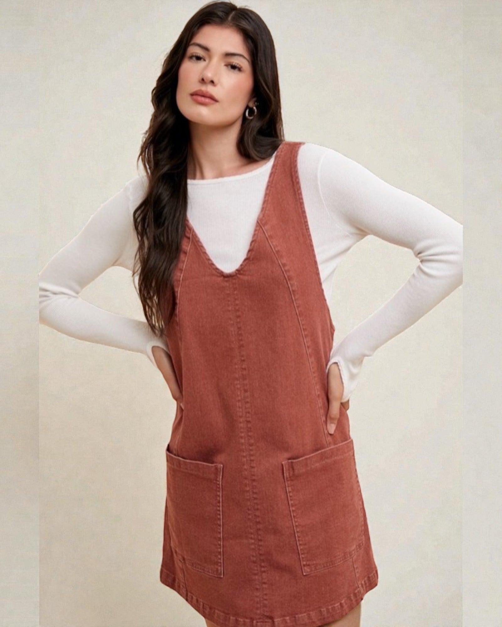 Woman wearing a brown pinafore dress over a white long-sleeve shirt on a beige background