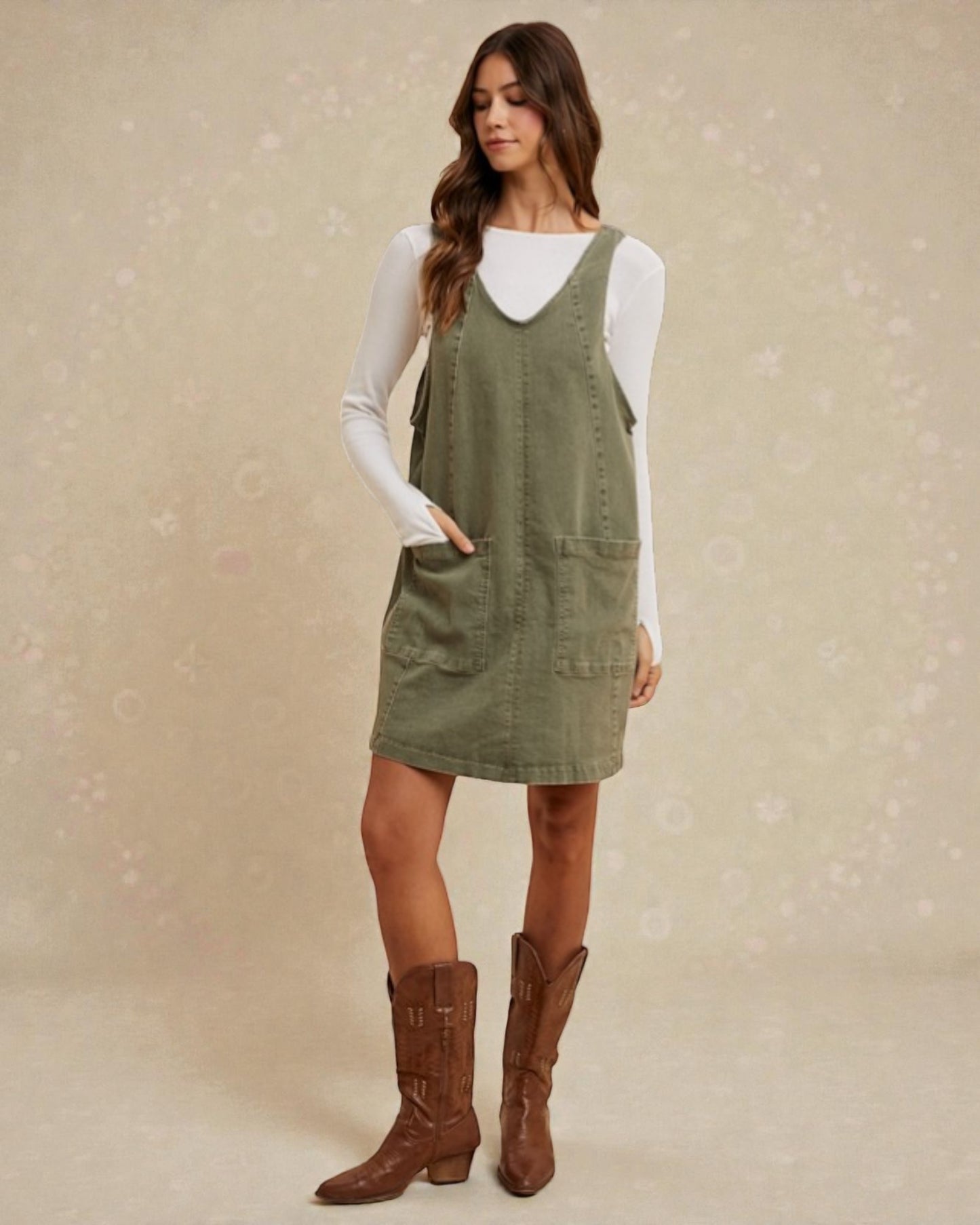 Woman wearing a green pinafore dress with a white long-sleeve shirt underneath, standing against a beige background.