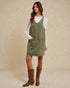 Woman wearing a green pinafore dress with a white long-sleeve shirt underneath, standing against a beige background.