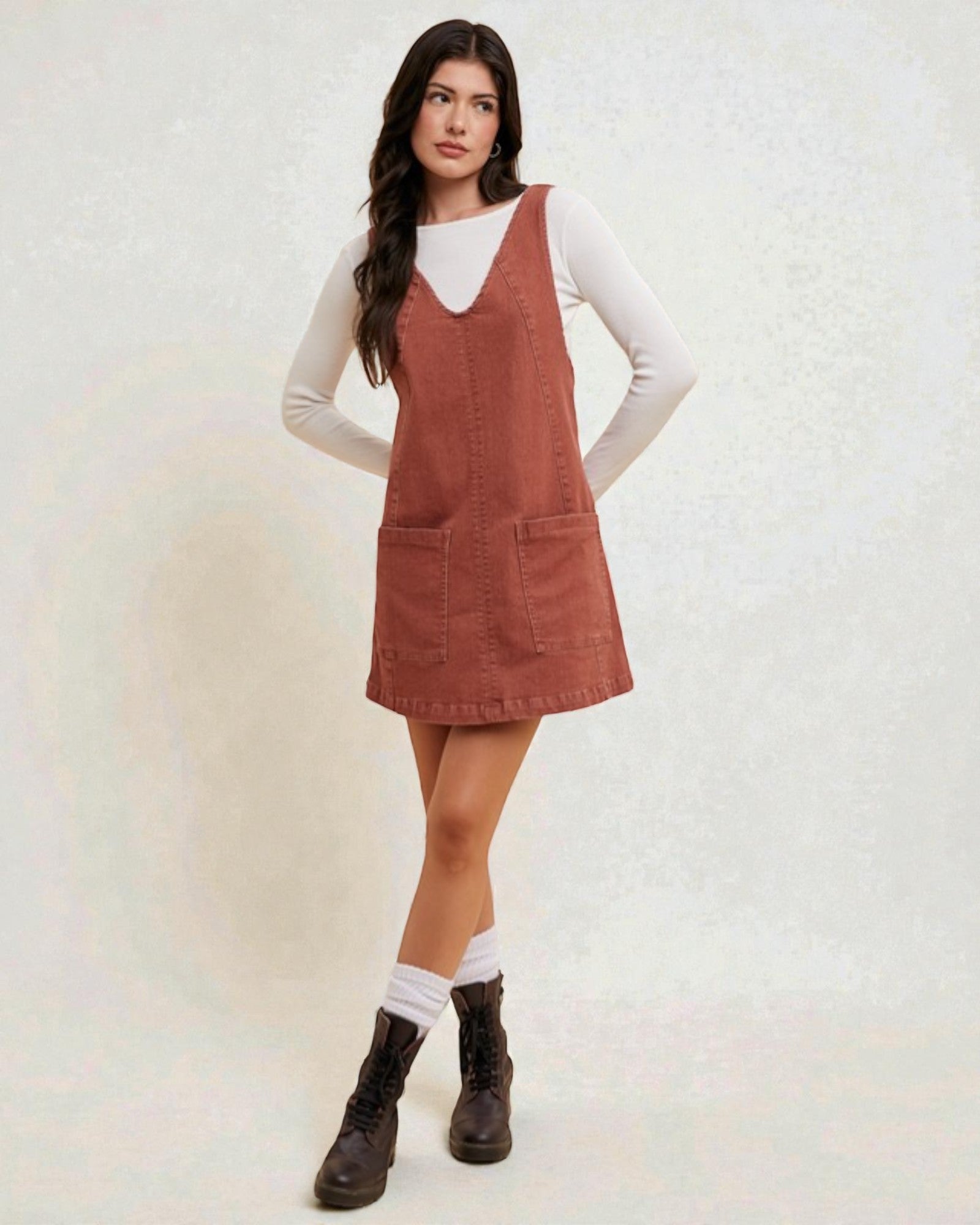 Woman wearing a brown pinafore dress over a white shirt on a light background
