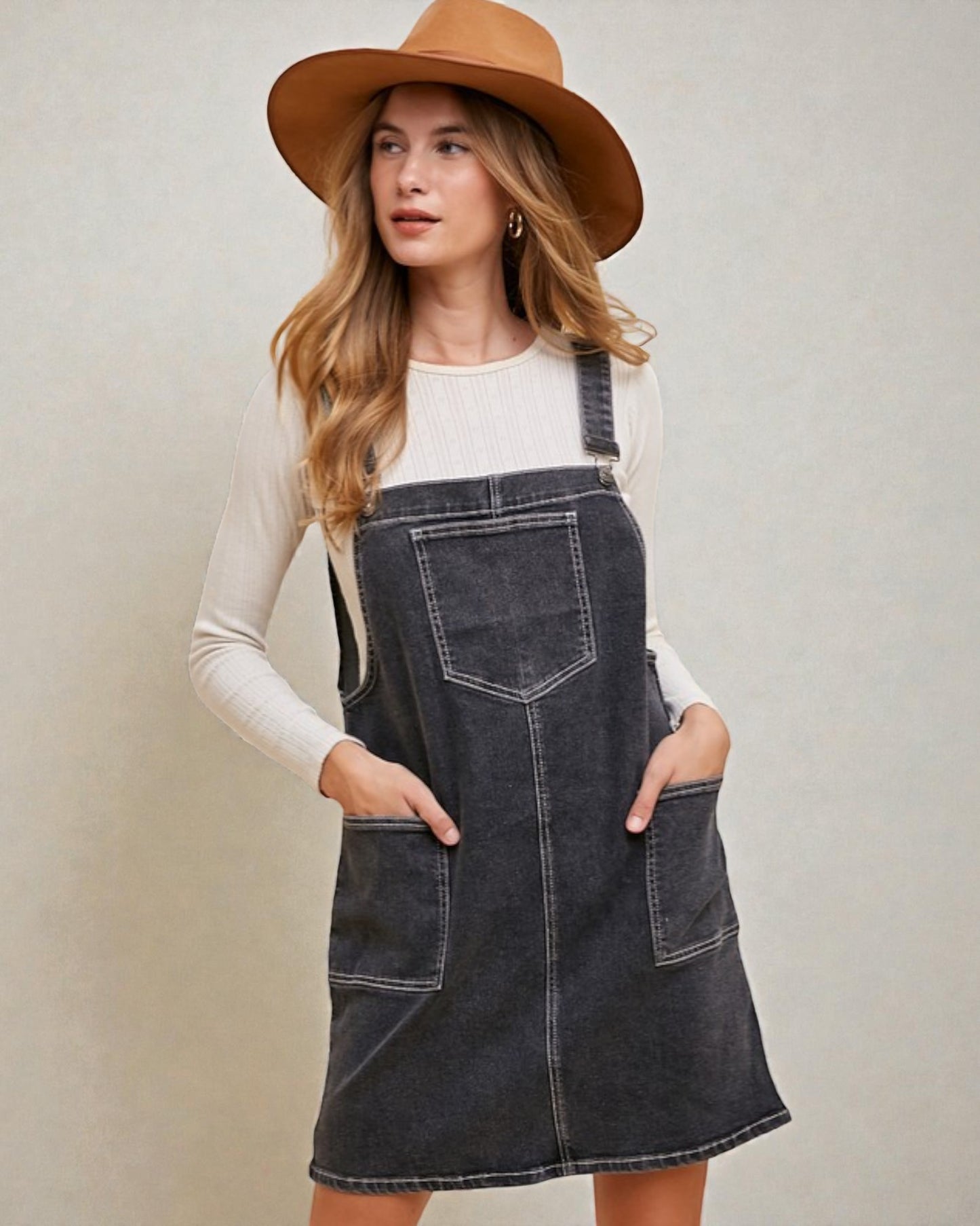Woman wearing a denim pinafore dress and brown hat against a plain background