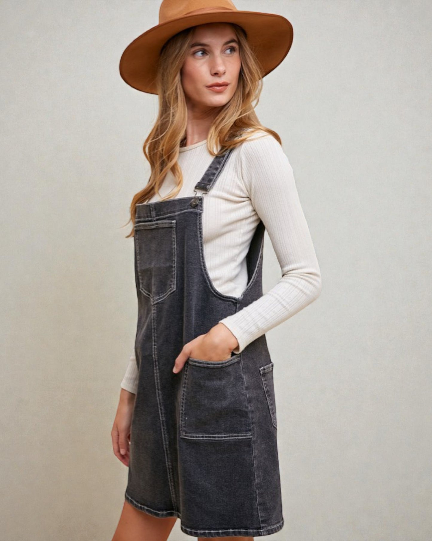 Woman wearing a denim pinafore dress and brown hat on a plain background