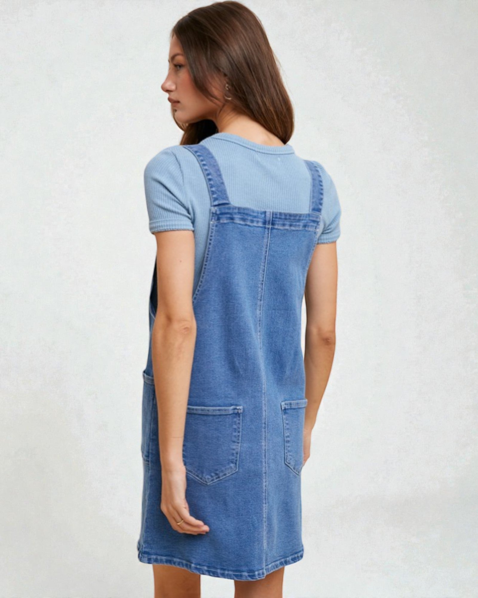 Woman wearing a blue denim pinafore dress over a light blue t-shirt on a white background