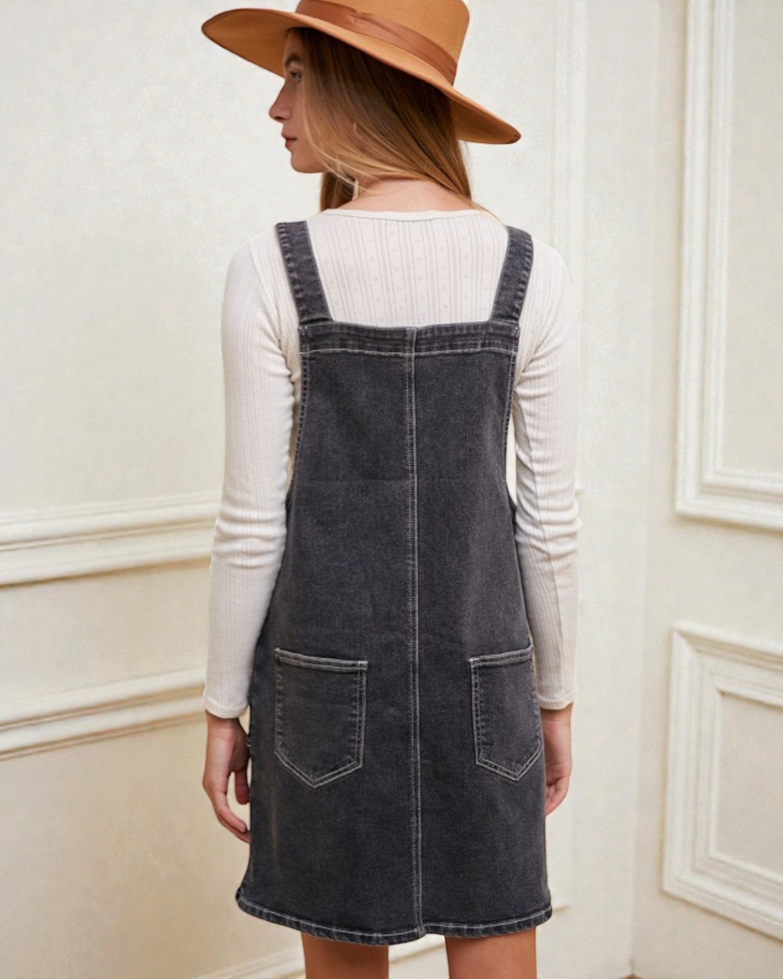 Woman wearing a denim pinafore dress and tan hat against a white wall.