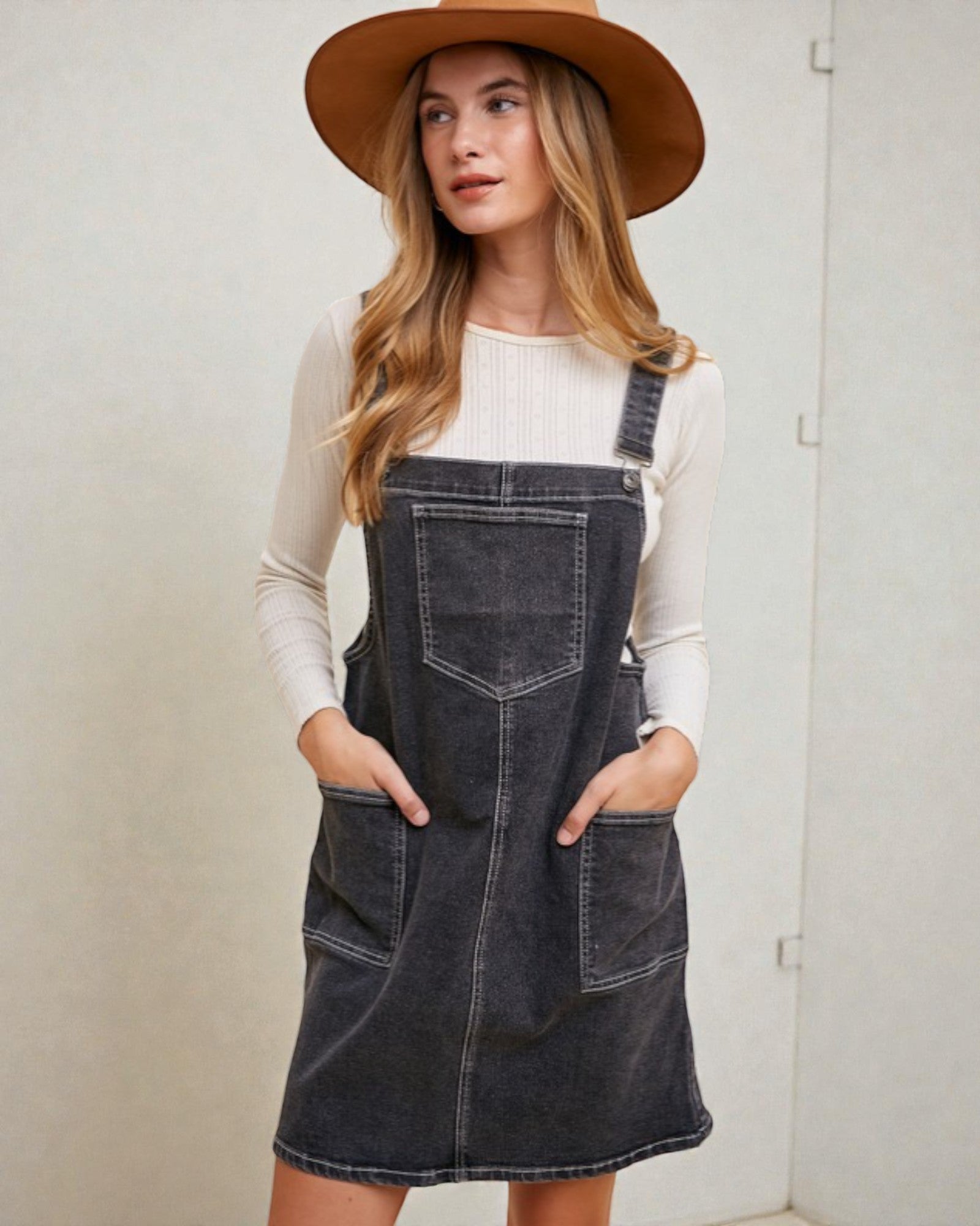 Woman wearing a denim pinafore dress and brown hat against a plain background
