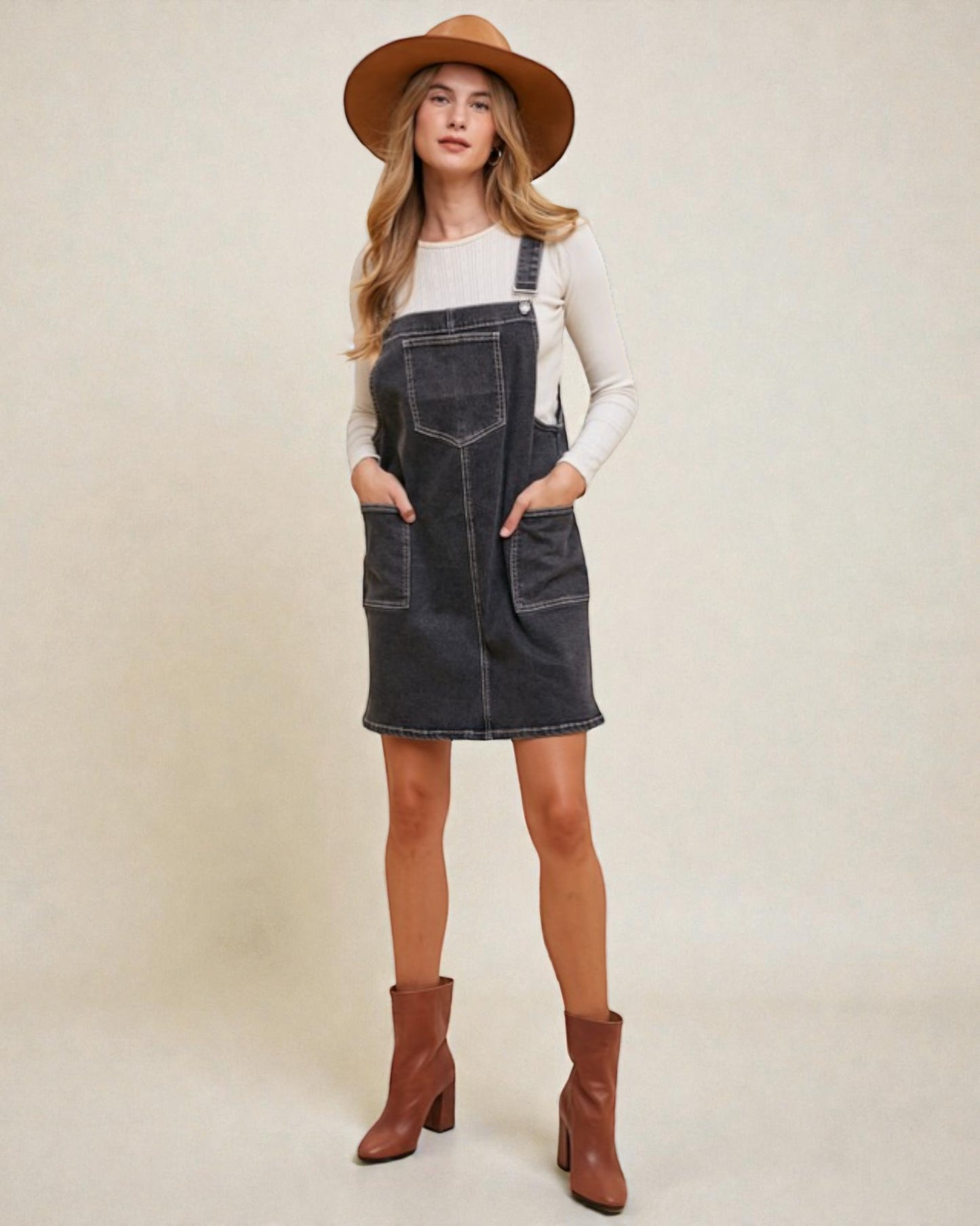 Woman wearing a denim overall dress with brown boots and a hat on a plain background