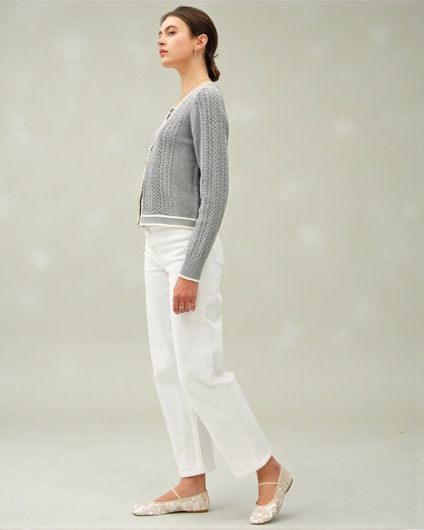 Woman wearing a gray cardigan, white pants, and flats on a plain background