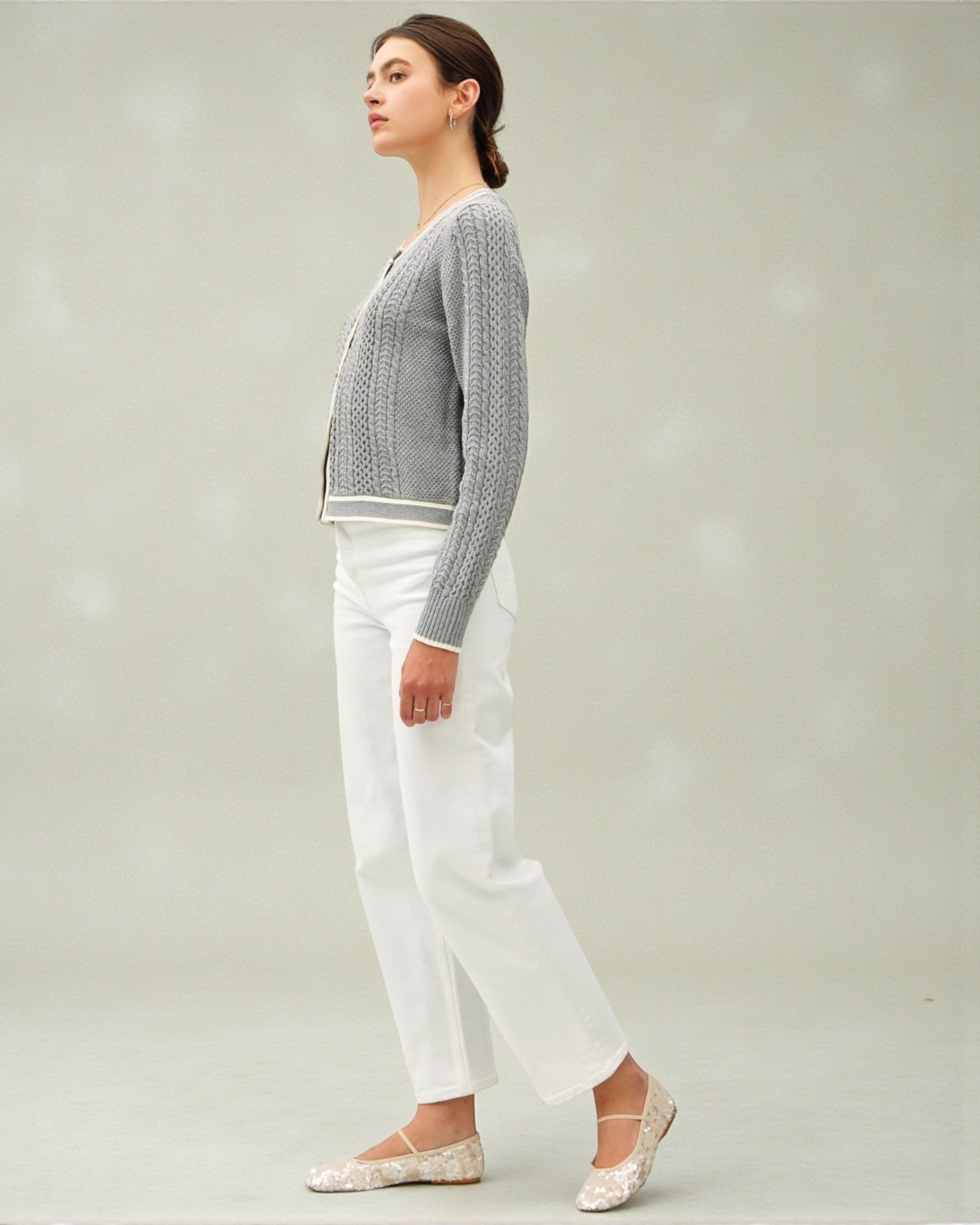 Woman wearing a gray cardigan, white pants, and flats on a plain background
