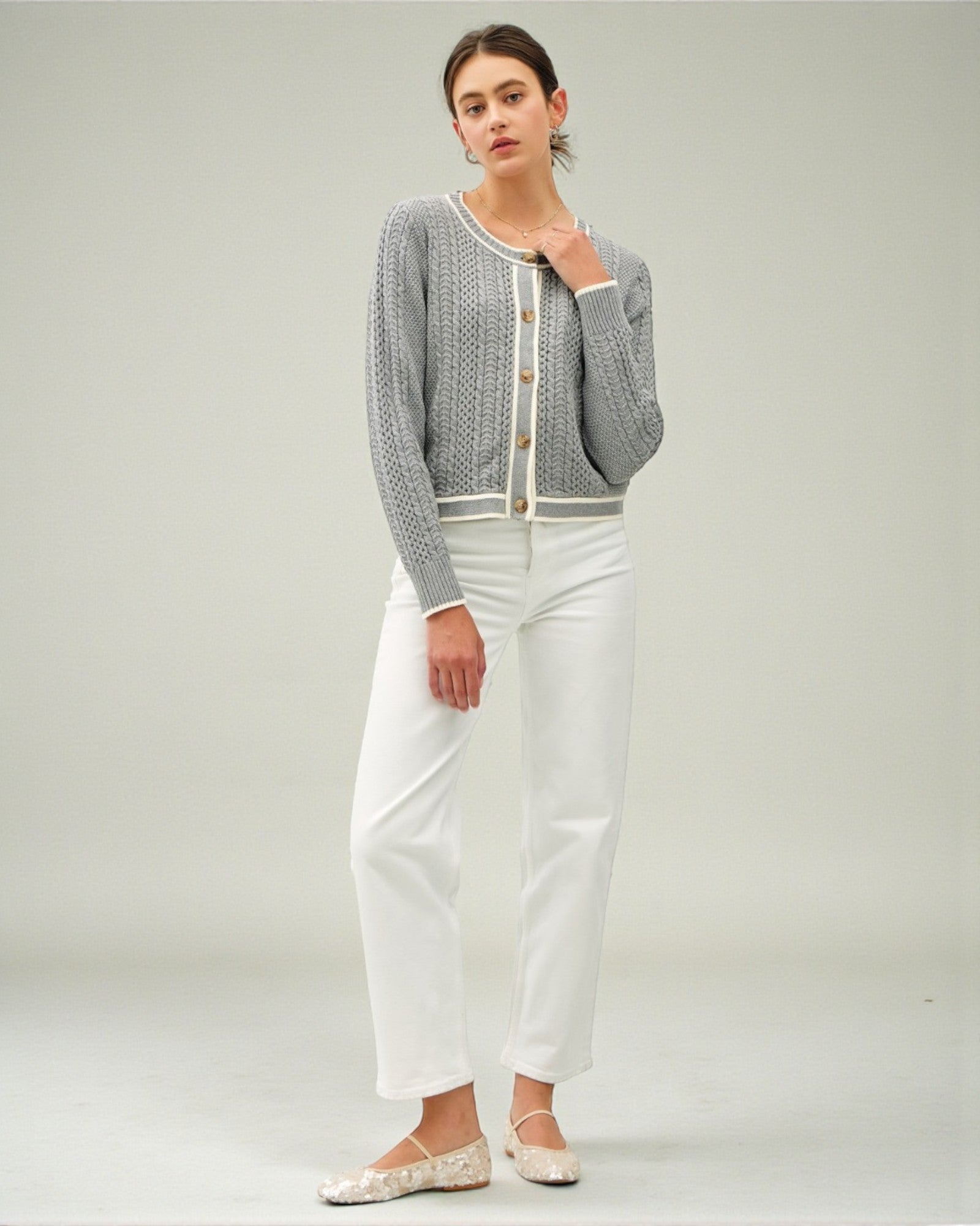Woman wearing a gray cardigan over a white top with white pants on a plain background
