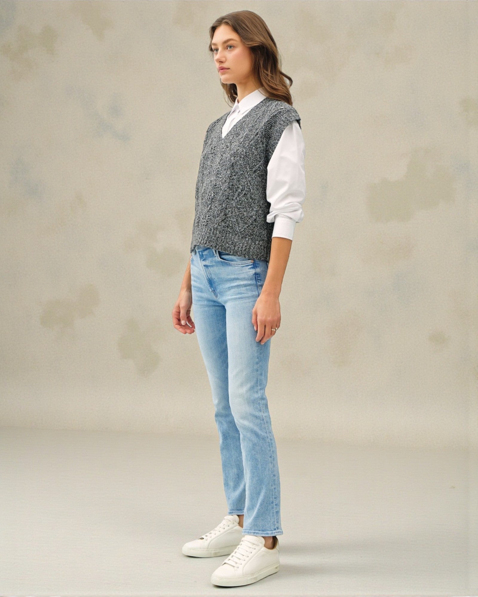 Woman wearing a gray knitted vest, white shirt, light blue jeans, and white sneakers on a neutral background