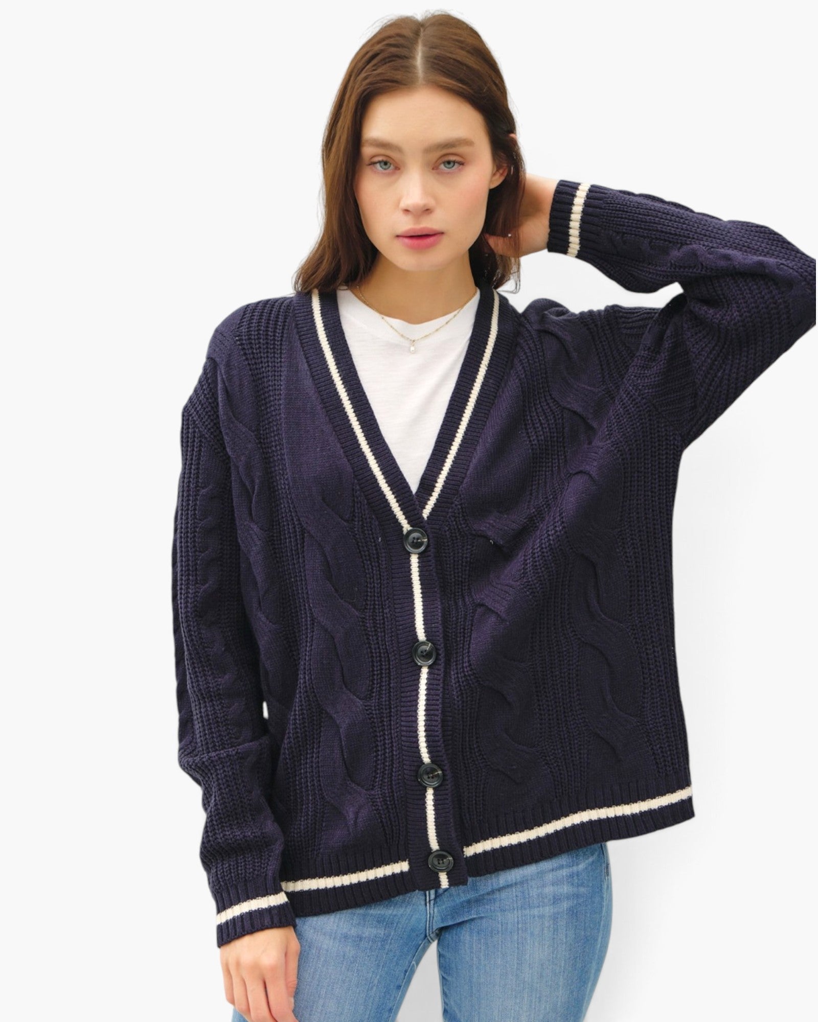 A woman modeling a dark purple cable-knit cardigan sweater with cream-colored trim and buttons, worn over a white t-shirt and blue jeans against a plain white background.