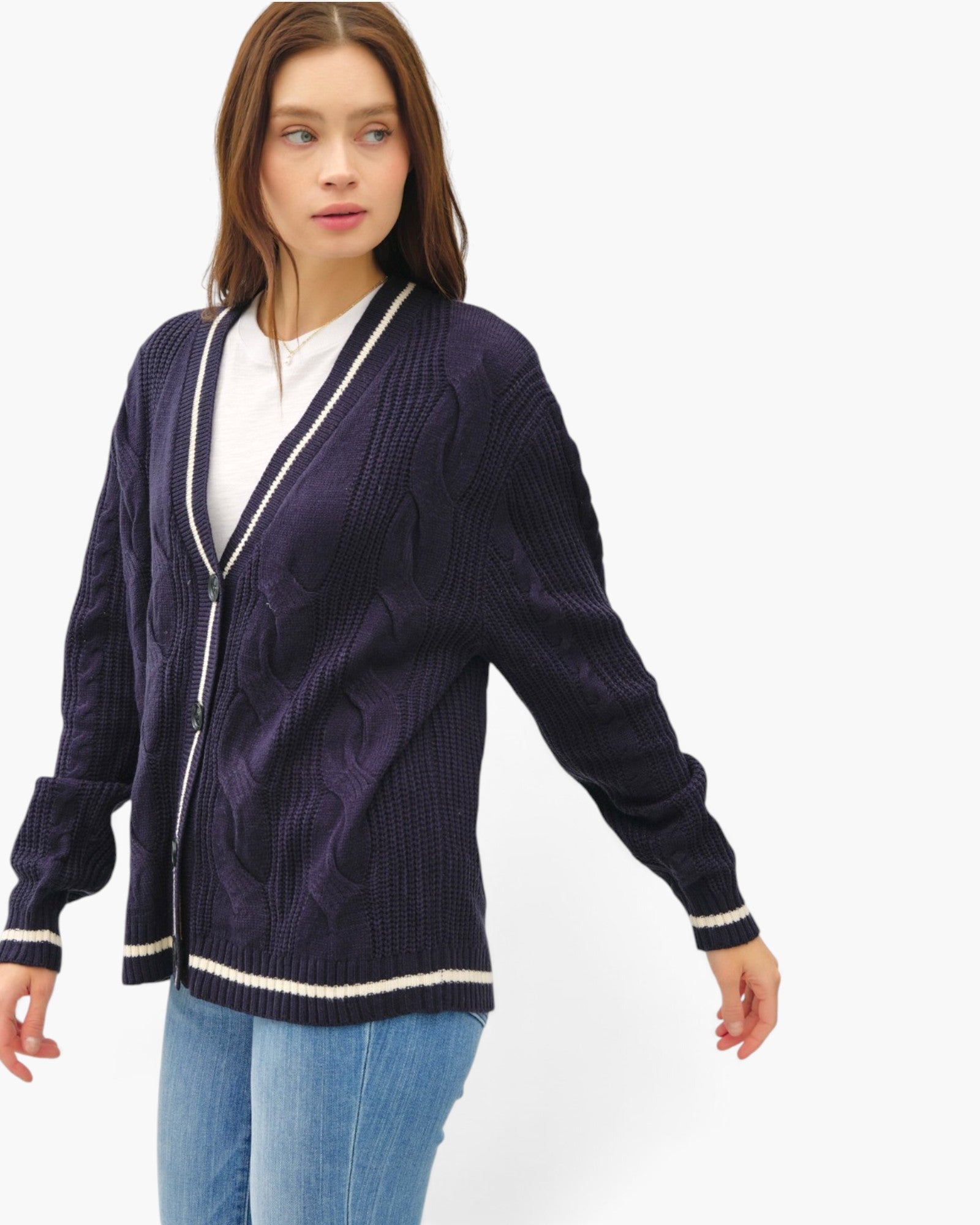 A woman wearing a dark purple cable-knit cardigan with cream trim over a white top and blue jeans, photographed against a plain light gray studio background.