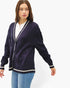 A woman wearing a dark purple cable-knit cardigan with cream trim over a white top and blue jeans, photographed against a plain light gray studio background.