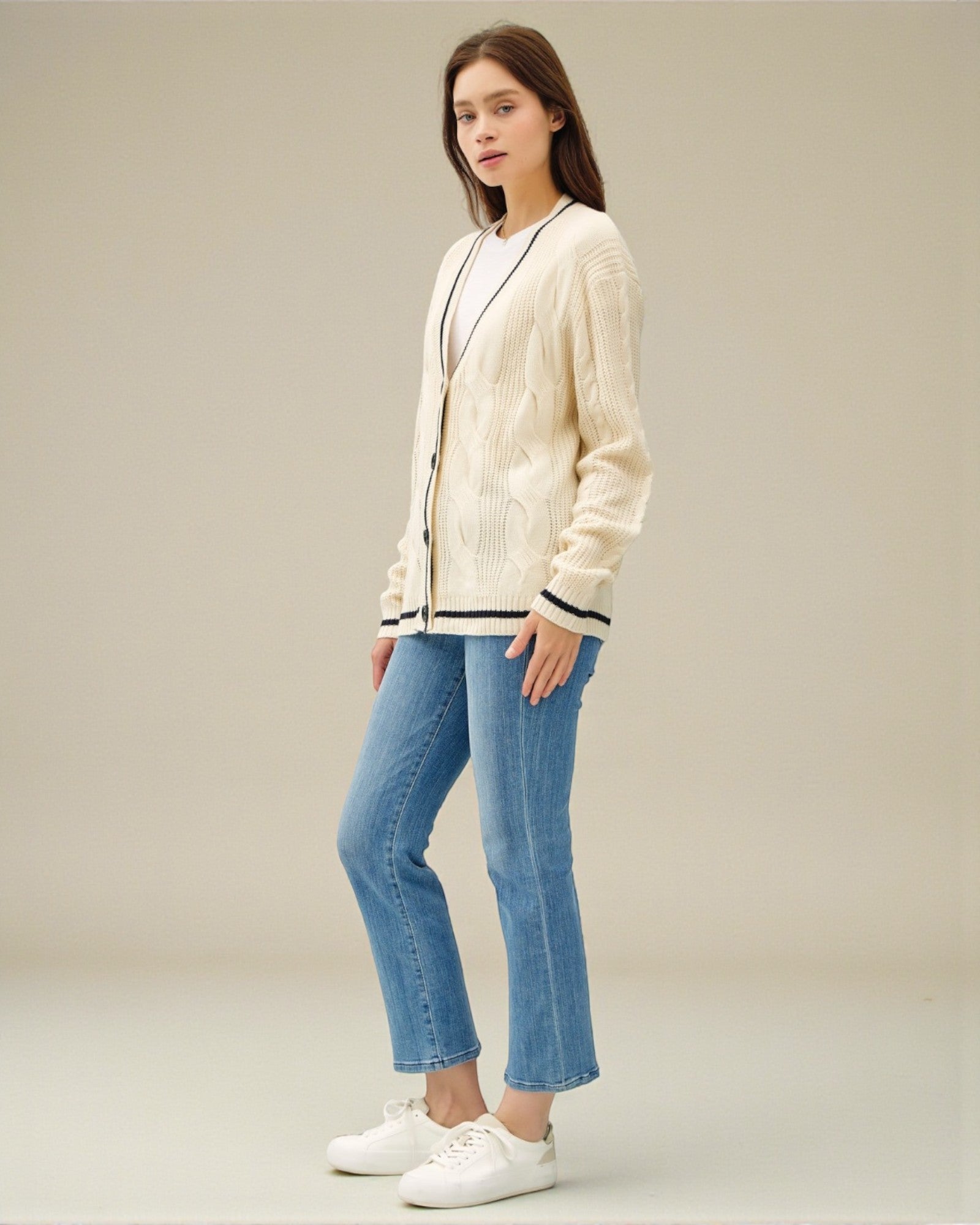 Woman wearing a cream cardigan with black trim, blue jeans, and white sneakers on a beige background