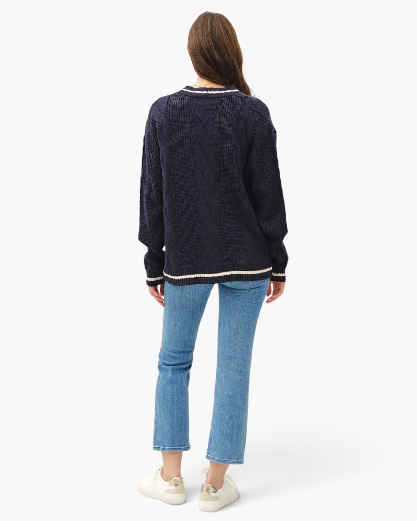 A woman shown from the back wearing a navy blue cable-knit sweater with cream ribbed trim, light blue cropped jeans, and white sneakers against a plain white studio background.