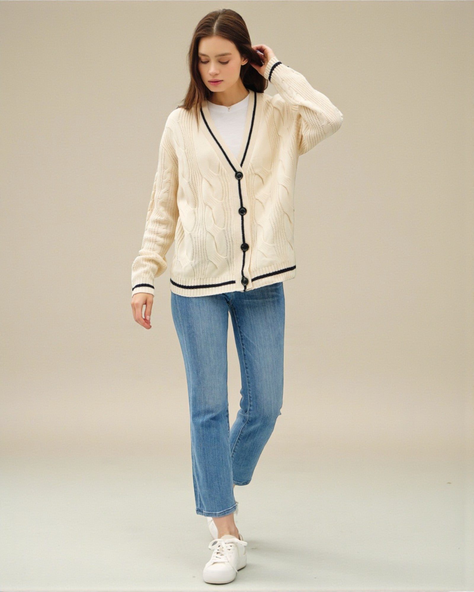 Woman wearing a cream cardigan with black buttons, white sneakers, and blue jeans on a beige background