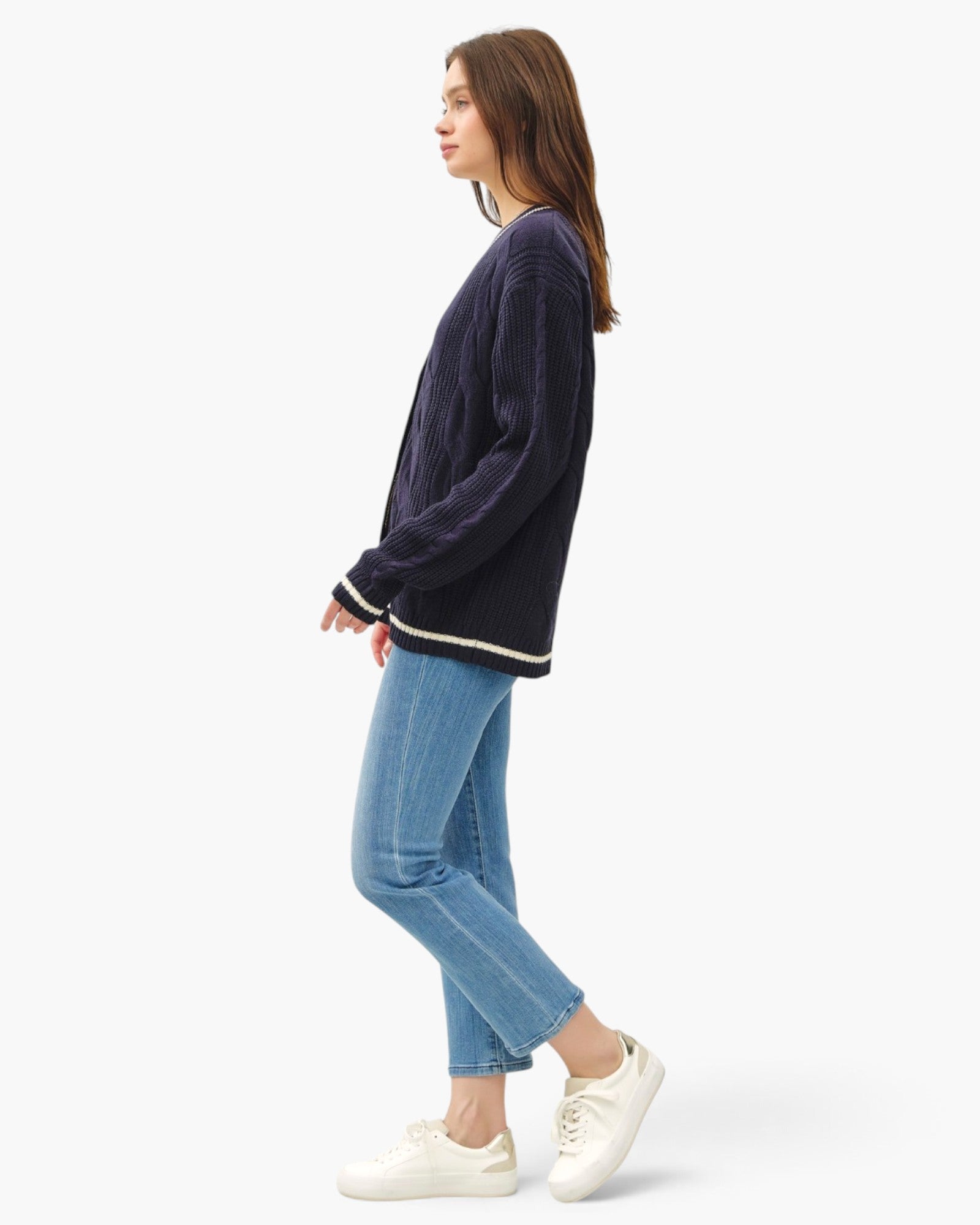 A woman in profile wearing a dark navy ribbed knit sweater with cream trim, blue cropped jeans, and white sneakers, photographed against a clean white studio background.