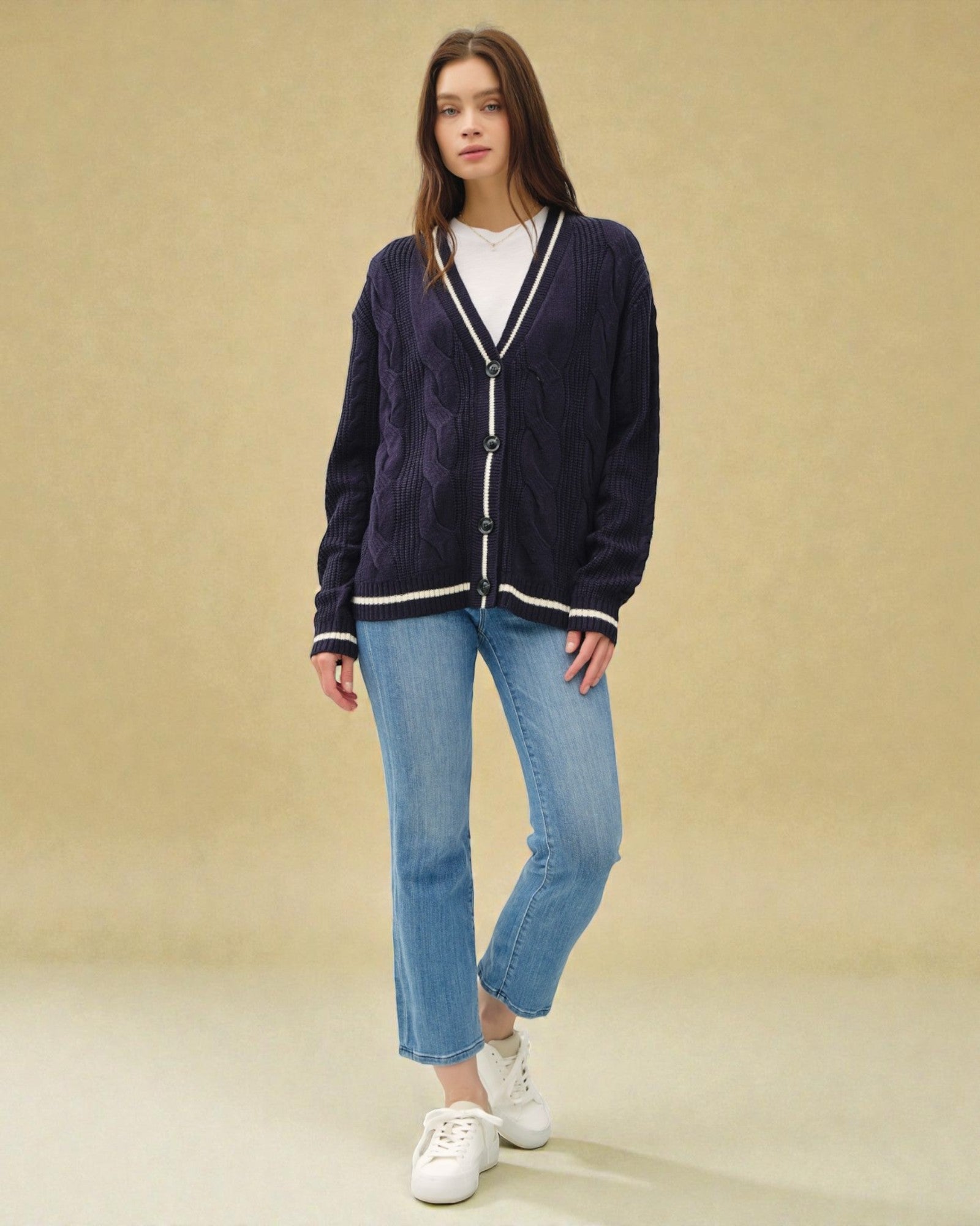 Woman wearing a navy cardigan, white shirt, blue jeans, and white sneakers on a beige background