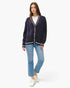 A woman modeling a dark purple cable-knit cardigan with white trim over a white t-shirt, blue jeans, and white sneakers, set against a plain white studio background.