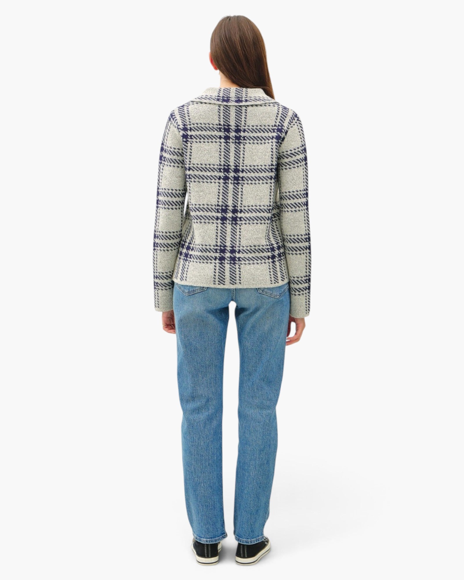 A woman viewed from the back wearing a cream-colored plaid blazer with navy and gray lines, paired with blue jeans and black sneakers, against a plain white studio background.
