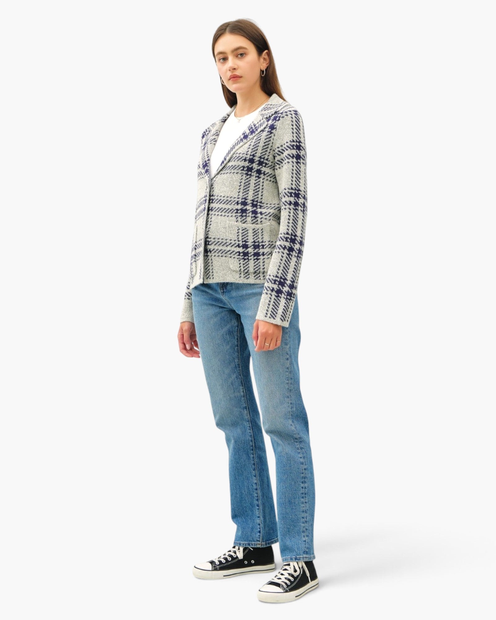 A woman modeling a white and navy plaid button-up cardigan over a white t-shirt, paired with blue jeans and black canvas sneakers, against a plain white studio background.